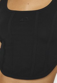 Gros plan d'un débardeur noir ajusté avec des détails de coutures verticales et un petit logo brodé en demi-cercle sur la poitrine.
