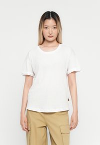 Woman with straight, shoulder-length hair wearing a plain white t-shirt and beige cargo pants, standing against a white background.