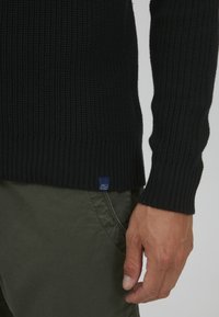 Schwarzer gerippter Pullover mit figurnaher Passform, mit einem sichtbaren blauen Etikett am Saum. Über olivgrünen Hosen getragen, die ein strukturiertes Strickmuster zeigen.