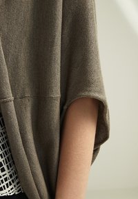 Brązowy dziergany kardigan z szerokimi rękawami i luźnym krojem. Nałożony na biały top z geometrycznym wzorem. Widoczna miękka faktura.