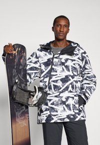 Σακάκι snowboard σε μαύρο και άσπρο με αφηρημένα σχέδια, διαθέτει μπροστινό φερμουάρ, κουκούλα και τσέπες, κρατώντας μια σανίδα snowboard και δέστρες.