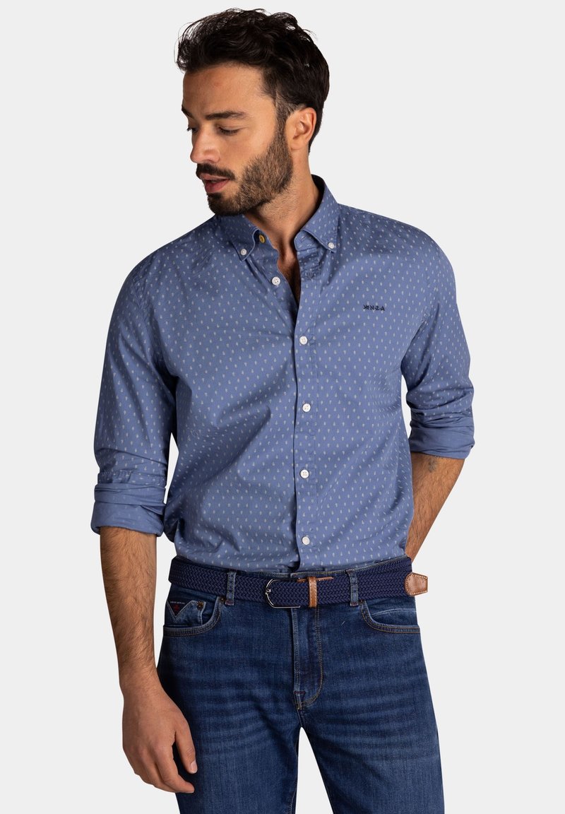 Blauw button-up overhemd met kleine witte geometrische patronen, opgerolde mouwen en een kraag. Draagt het met donkere spijkerhosen en een gevlochten marineblauwe riem.
