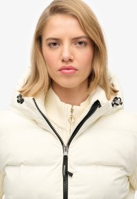Biała kurtka puchowa z kapturem, z czarnym zamkiem i uchwytem. Nosi się ją na kremowym swetrze ribbed, który ma gładką fakturę.