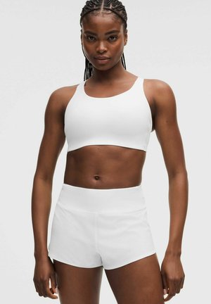 Frau mit geflochtenem Haar, die einen weißen Sport-BH und weiße Sportshorts trägt, steht vor einem schlichten hellen Hintergrund.
