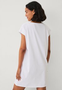 Hvid bomulds T-shirt kjole med korte ærmer, afslappet pasform, rund halsudskæring og slidser i siderne for nem bevægelse. Glat, blød tekstur.