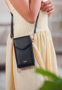 Černá kožená křížová taška se zlatým kováním, zapínáním na zip a zakřiveným klopovým víkem. Na tašce se nachází logo v růžové barvě, která je nošena k světle žlutým šatům.