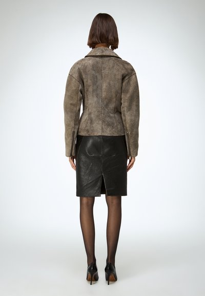 Barna szarvasbőr dzseki, bemetszett gallérral, oldalsó cipzárokkal és testhez simuló szabással, fekete bőrszoknyával párosítva, amely hátul hasítékkal rendelkezik.