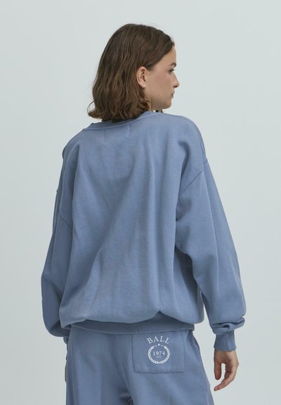 Liels zils svīteris ar ribotām piedurknēm un apakšmalu, ar mazu baltu logo aizmugurē. Izgatavots no mīksta, samtu auduma.