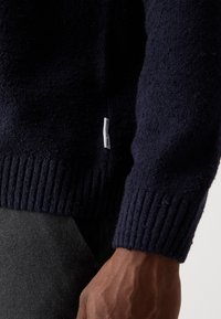 Navy knitted sweater with ribbed cuffs, made of textured fabric. Features a small label on the side, shown worn over dark gray pants.