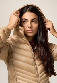 Beige pufferjack met een capuchon, met horizontale gestikte naden, een rits aan de voorkant en twee zijzakken. Gladde, glanzende textuur.