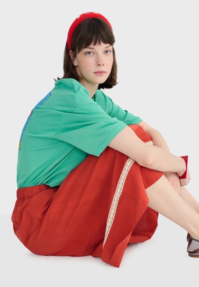 Jeune femme aux cheveux bruns courts portant un t-shirt vert, une jupe rouge avec de la dentelle, un bandeau rouge, assise en tailleur sur un fond blanc.