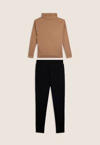 Maglia a maniche lunghe marrone con collo alto abbinata a pantaloni aderenti neri su uno sfondo chiaro e uniforme.