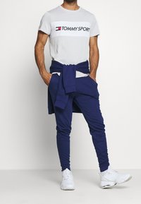 Conjunto masculino que incluye una camiseta gris con un panel blanco y la marca "TOMMY SPORT", combinada con joggers azul marino y zapatillas deportivas blancas.