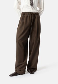 Bruine broek met wijde pijpen, elastische tailleband en trekkoord, gemaakt van gladde stof; gecombineerd met donkere schoenen en een licht shirt.