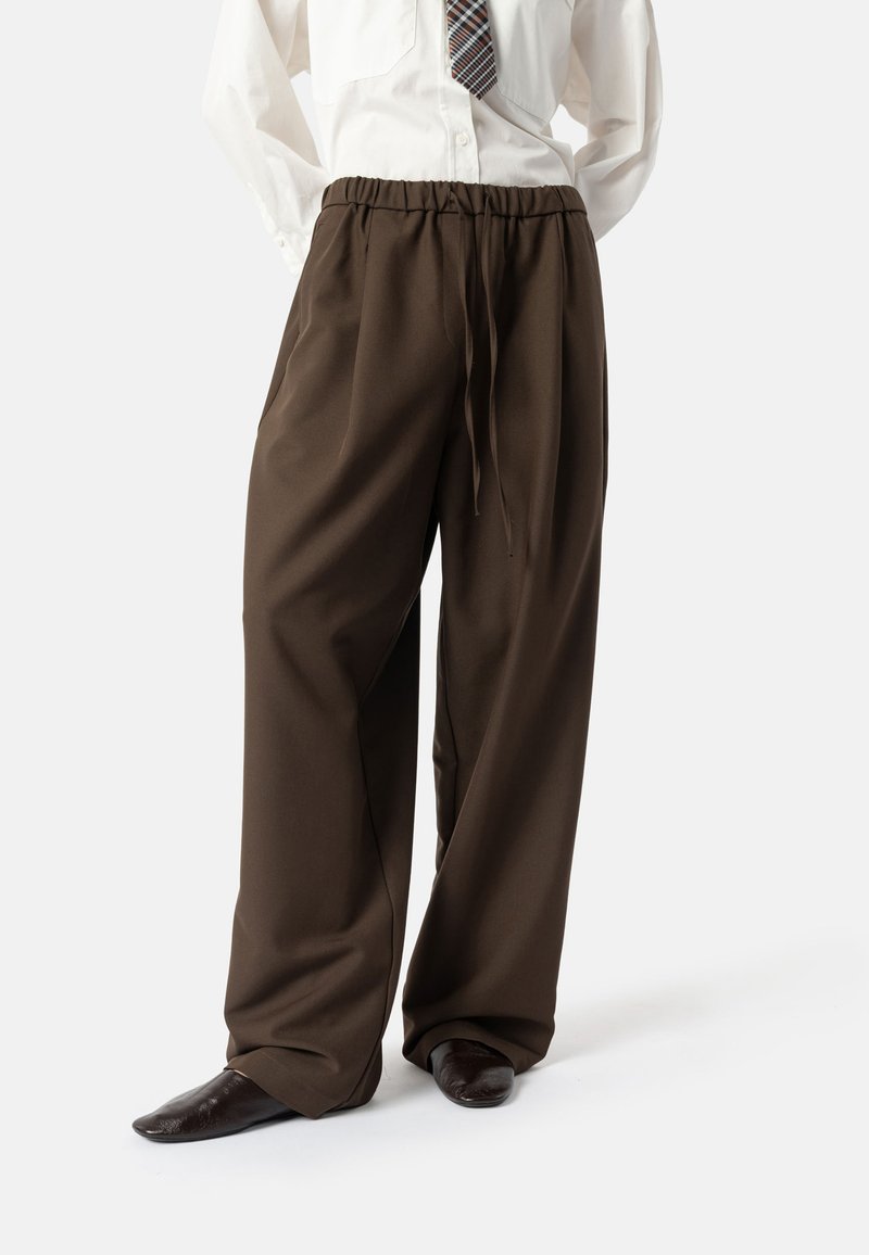 Bruine broek met wijde pijpen, elastische tailleband en trekkoord, gemaakt van gladde stof; gecombineerd met donkere schoenen en een licht shirt.