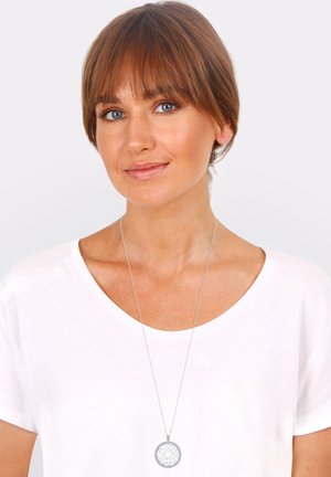Silberner Anhänger mit kunstvollem kreisförmigem Design, auf einer dünnen Kette aufgehängt. Getragen über einem schlichten weißen T-Shirt.
