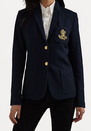 Femme portant un blazer bleu marine ajusté avec des boutons dorés et un écusson brodé sur la poche poitrine, par-dessus une chemise blanche boutonnée.