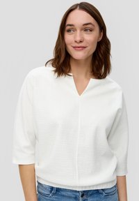 Weißes, strukturiertes Blusenshirt mit V-Ausschnitt, Dreiviertelärmeln und lockerem Schnitt, kombiniert mit blauen Denim-Jeans. Zarte Musterdetails sichtbar.