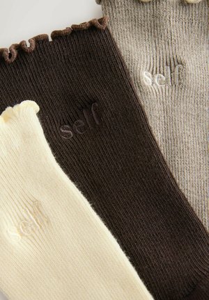 Drie paar geribde sokken: crème met een gerimpelde rand, donkerbruin met subtiel logo, en beige met een gestreepte textuur.