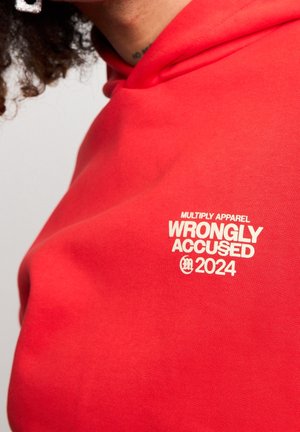 Czerwona bluza z kapturem z białym napisem na piersi "MULTIPLY APPAREL WRONGLY ACCUSED 2024", noszona przez osobę z kręconymi włosami.