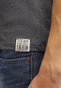 Grå bomuldsskjorte med et hvidt mærkat, der står "STEAM AND DRY IRON TOM TAILOR." Bæres med blå denim jeans med en kobberknap.
