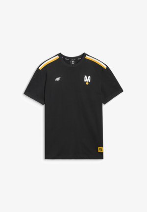 Mężczyzna z krótkimi blond włosami nosi czarną koszulkę z żółtymi i białymi pasami na ramionach oraz logo, stoi na tle jednolitego jasnego tła.