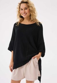 Czarny dzianinowy sweter z szerokim dekoltem, rękawami o długości trzech czwartych oraz asymetrycznym dołem, w zestawieniu z jasnobeżowymi szortami. Gładka tekstura.