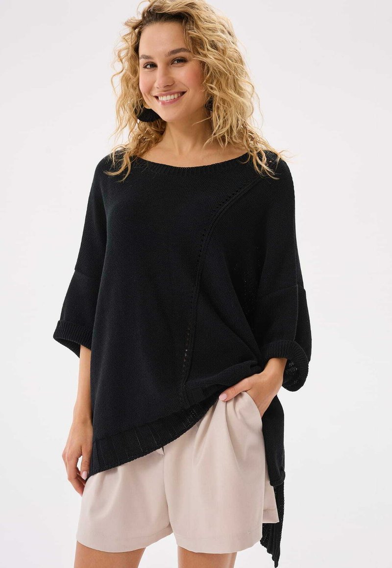 Czarny dzianinowy sweter z szerokim dekoltem, rękawami o długości trzech czwartych oraz asymetrycznym dołem, w zestawieniu z jasnobeżowymi szortami. Gładka tekstura.
