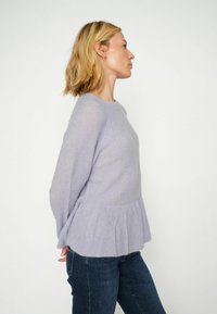 Svetlo sivoljubičasti pulover z žametsko teksturo, okroglo izrezom in peplum robom. Dolgi rokavi in sproščen kroj, oblečen preko temno modrih kavbojk.