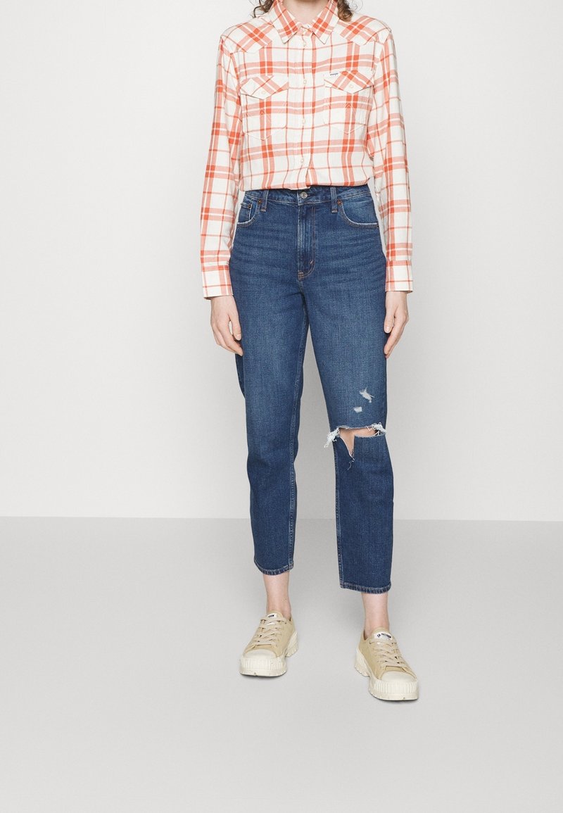 Camisa de cuadros de manga larga en naranja y blanco, combinada con jeans de talle alto azul con detalles desgastados en una rodilla y zapatillas beige.