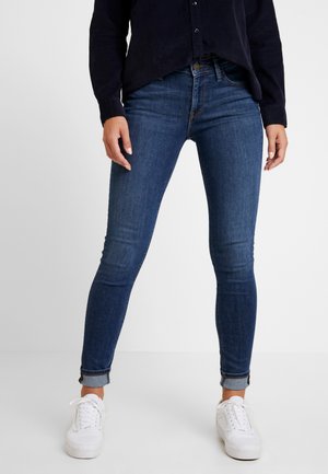 Personne portant un jean skinny bleu foncé retroussé aux chevilles, des baskets blanches et un t-shirt noir à manches longues, debout devant un fond uni.