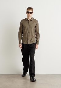 Camisa de manga longa, verde-azeitona, com design de botão, combinada com calças pretas e óculos de sol, contra um fundo liso.