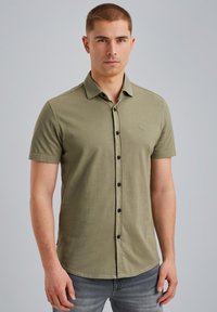 Kratki rukavi maslinasto zeleni slim fit košulja s rebrastom teksturom i crnim gumbima, uz suptilan logo na prsima, u kombinaciji s sivim trapericama.
