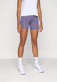 Lilla sportskort, lavet af strækbart materiale, med en tætsiddende design og kontrastsyet. Båret med en hvid top og sportssko.