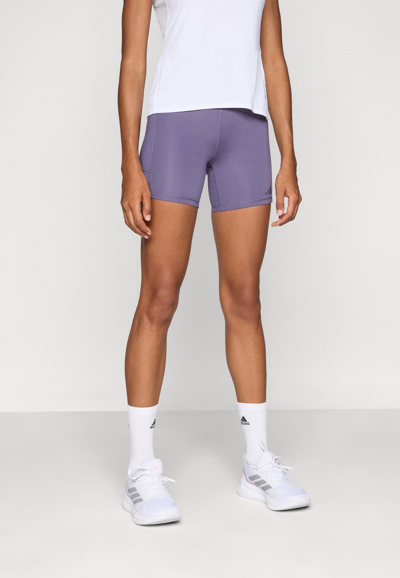 Lilla sportskort, lavet af strækbart materiale, med en tætsiddende design og kontrastsyet. Båret med en hvid top og sportssko.