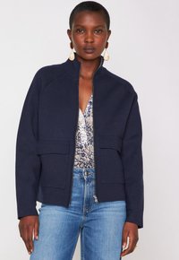 Donna che indossa una giacca blu navy con zip e tasche a pattina sopra una camicia floreale con bottoni e jeans azzurri chiari, accessoriate con orecchini a goccia.
