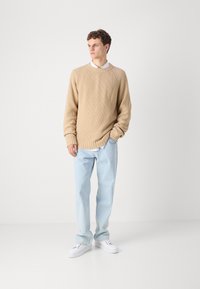 Beige ribbed knit sweater with a crew neckline, paired with light blue straight-leg jeans and white sneakers, set against a plain background.