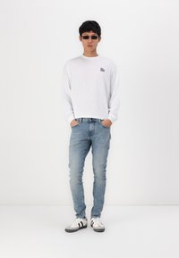 White long-sleeve shirt with small black text, light blue jeans, and white sneakers with black accents. Model wearing black sunglasses.