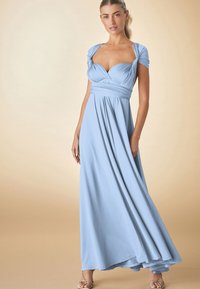 Mujer de pie vistiendo un vestido largo hasta el suelo de color azul claro con mangas cortas retorcidas y un corpiño fruncido sobre un fondo neutro.