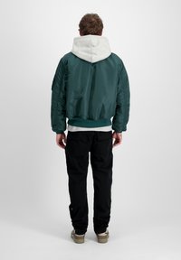 Zelená bomber bunda s šedou mikinou, žebrovanými manžetami a lemom, skombinovaná s čiernymi nohavicami. Bunda má lesklý, hladký povrch.