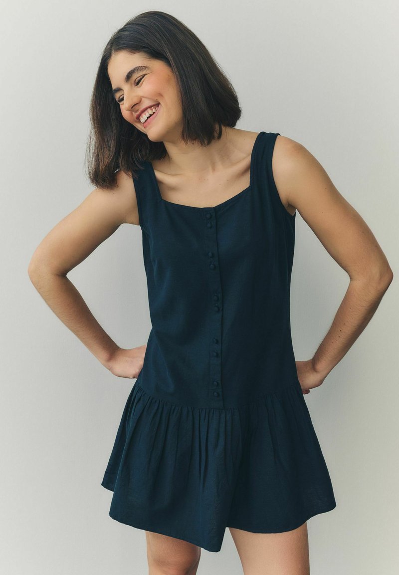 Rochie fără mâneci, de culoare bleumarin, cu decolteu pătrat, deschidere cu nasturi și fustă evazată. Materialul pare moale, cu o ușoară textură.