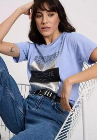 Jasnoniebieska koszulka z nadrukiem w kolorze srebrnym przedstawiającym wzorzysty prostokąt oraz tekst "WIERZ w siebie", w połączeniu z dżinsami z wysokim stanem.