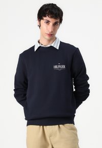 Marineblauwe sweatshirt met een ronde hals, voorzien van een klein wit "HILFIGER" logo. Draagt over een gestreept overhemd met kraag. Zachte stof, casual ontwerp.