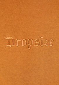 Tissu en coton de couleur orange brûlé présentant le mot "Dropsize" brodé dans une police de style gothique, mettant en avant la texture et le savoir-faire.