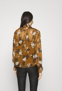 Blouse fleurie marron à manches longues, avec une texture transparente, un col froncé et des motifs de feuilles noires sur fond crème.