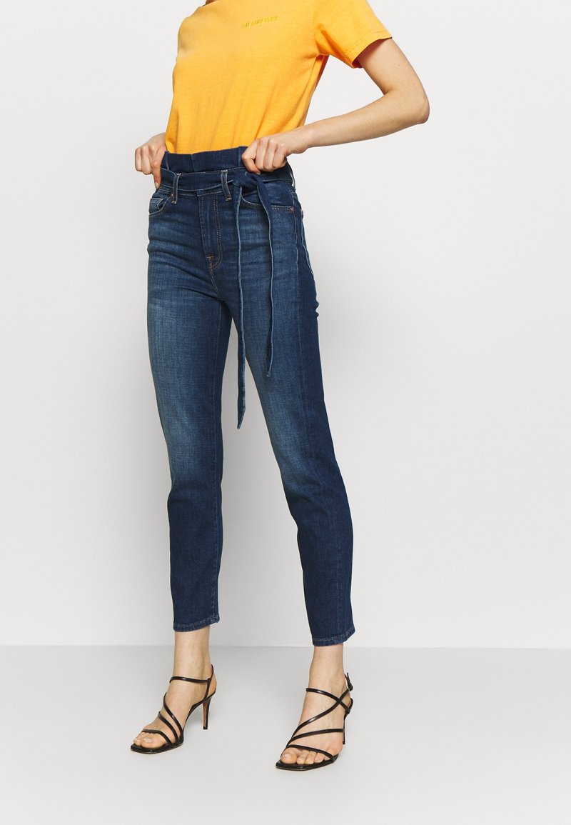 Jeans de tiro alto en azul oscuro con un diseño de cinturón, que cuentan con un corte ajustado y largo hasta los tobillos, combinados con sandalias de tacón alto negras con tiras.