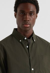Uomo con capelli ricci corti che indossa una camicia verde oliva con bottoni e tasca, rivolto in avanti su uno sfondo chiaro uniforme.