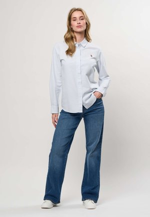 Tânără stând cu o mână în buzunar, purtând o cămașă cu dungi albastre deschis cu nasturi, blugi albaștri și teniși albi pe fundal simplu.