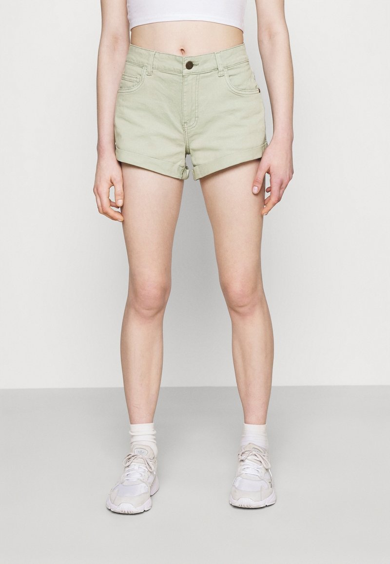 Vaaleanvihreät farkkushortsit, joissa on korkea vyötärö, käännetyt lahkeet, etutaskut ja yksi nappisulku. Yhdistetty valkoisiin lenkkareihin ja sukkiin.