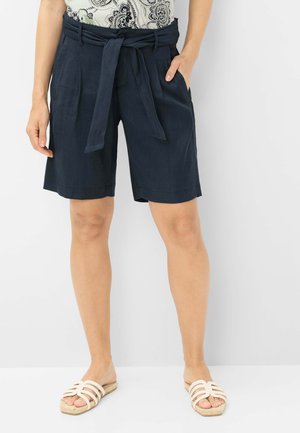 Femme portant un short taille paperbag bleu marine avec ceinture nouée, un top à motifs et des sandales blanches à enfiler, debout les mains dans les poches.